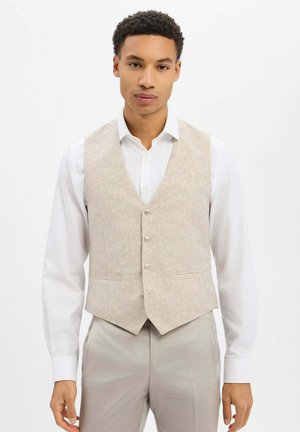 Beige Weste mit strukturiertem Design, mit V-Ausschnitt, sechs Knöpfen, zwei Taschen vorne, getragen über einem weißen Hemd.