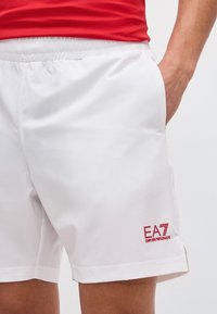 Άνδρας που φοράει λευκά σορτς EA7 Emporio Armani και κόκκινο πουκάμισο, με το χέρι στην τσέπη των σορτς, μπροστά σε απλό φόντο.