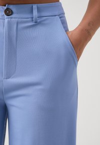 Detailní záběr na světle modré kalhoty s hnědým knoflíkem a ruku odpočívající v pravé kapse, na zápěstí je vidět malý tetování.