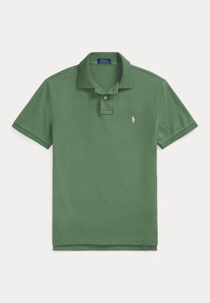 Homme portant un polo vert à manches courtes, un pantalon plissé crème, des mocassins marron à glands et une montre-bracelet, debout devant un fond beige uni.