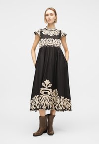 Γυναίκα στέκεται φορώντας μαύρο μίντι φόρεμα με μπεζ λουλουδένινη κεντητή διακόσμηση και καφέ μποτάκια αστραγάλου, με τα χέρια στις τσέπες, ουδέτερο φόντο.