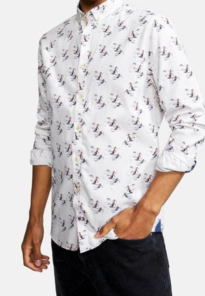 Man draagt een wit overhemd met knopen met een kleurrijke print van een skiër en een zwarte broek, één hand in de zak, mouwen opgerold.