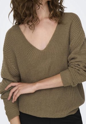 Femme portant un pull en tricot marron, ample et texturé, avec un col en V profond et des manches longues, un bras croisé sur l'autre au niveau de la taille.