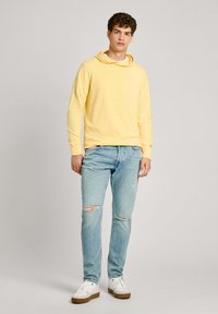 Sudadera amarilla con capucha y mangas largas, combinada con jeans de mezclilla claros y desgastados y zapatillas blancas con suelas marrones. Diseño simple, ajuste casual.