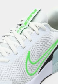 Hvit treningssko med teksturert mesh-overdel, grønne detaljer og fremtredende Nike swoosh-logo. Har flate lisser og polstret pløse.