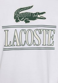 Camiseta blanca de algodón con un logo de cocodrilo bordado en verde sobre el texto "LACOSTE" en una fuente verde en negrita, enmarcada por dos líneas.