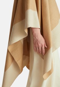 Pruhovaný pončo v odstínech tan a krémové barvy s měkkou texturou, volně zavěšenou látkou a vlnitým lemem. Ruka s více prsteny drží látku.