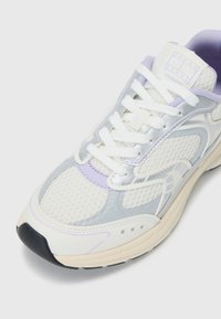 Sneaker bianco con sovrapposizioni in mesh e sintetico, caratterizzata da dettagli in argento e lavanda, motivi testurizzati e chiusura con lacci bianchi.