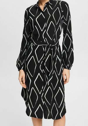 Robe noire boutonnée avec manches longues, motif losanges blancs et ceinture en tissu nouée à la taille, portée par une personne debout.