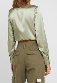Kvinna i en ljusgrön satincroppad blus och hög midja olivfärgade cargo byxor med knappade luckor, sedd bakifrån.