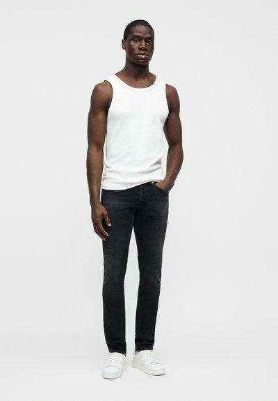 Άνδρας που στέκεται φορώντας λευκό αμάνικο φανελάκι, μαύρο στενό τζιν και λευκά αθλητικά παπούτσια, μπροστά σε απλό ανοιχτόχρωμο φόντο.