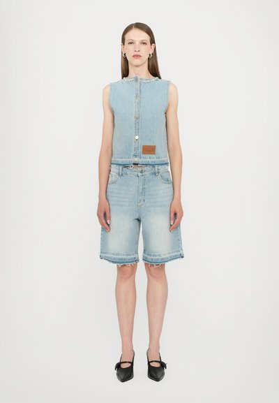 Denim odijelo s bezrukavom svijetloplavom prslukom s dugmadi i šortsevim. Šorts imaju izlizani rub i džepove. Upareno s crnim cipelama.