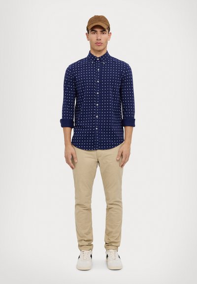 Jūrų jėgų marškiniai su baltais taškeliais, sukabinti rankovės, smėliniai chinos ir baltos sportbačiai su juodais akcentais; dėvimi su ruda kepure.