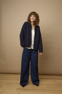 Femme aux cheveux bouclés, debout en souriant, portant un cardigan sombre boutonné, un top en dentelle blanche, un pantalon large bleu foncé et des chaussures pointues.