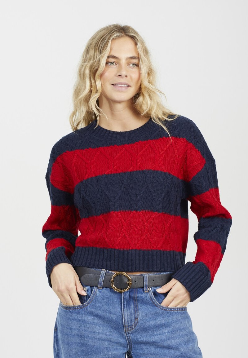 Pull en tricot rayé rouge et marine avec motif en câble, col rond, poignets et ourlet côtelés, associé à un jean bleu taille haute et une ceinture.