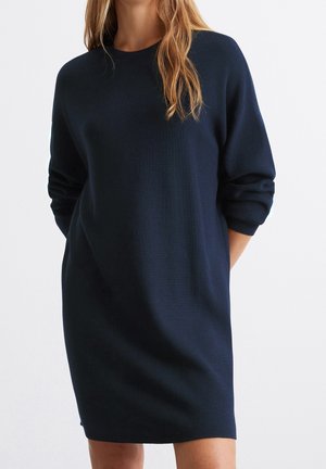 Vrouw draagt een marineblauwe, gebreide jurk met lange mouwen en een ronde hals, staand met haar handen achter haar rug.