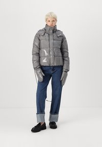 Grå pufferjakke med en shiny finish, festet hette, frontglidelås og store, puffy seksjoner; parret med blå jeans og hansker.