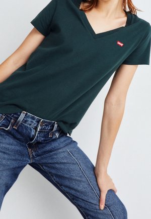 Henkilö, jolla on päällään tumma vihreä V-kauluksinen Levi's t-paita ja siniset farkut, käsi lepää reisillä yksiväristä taustaa vasten.