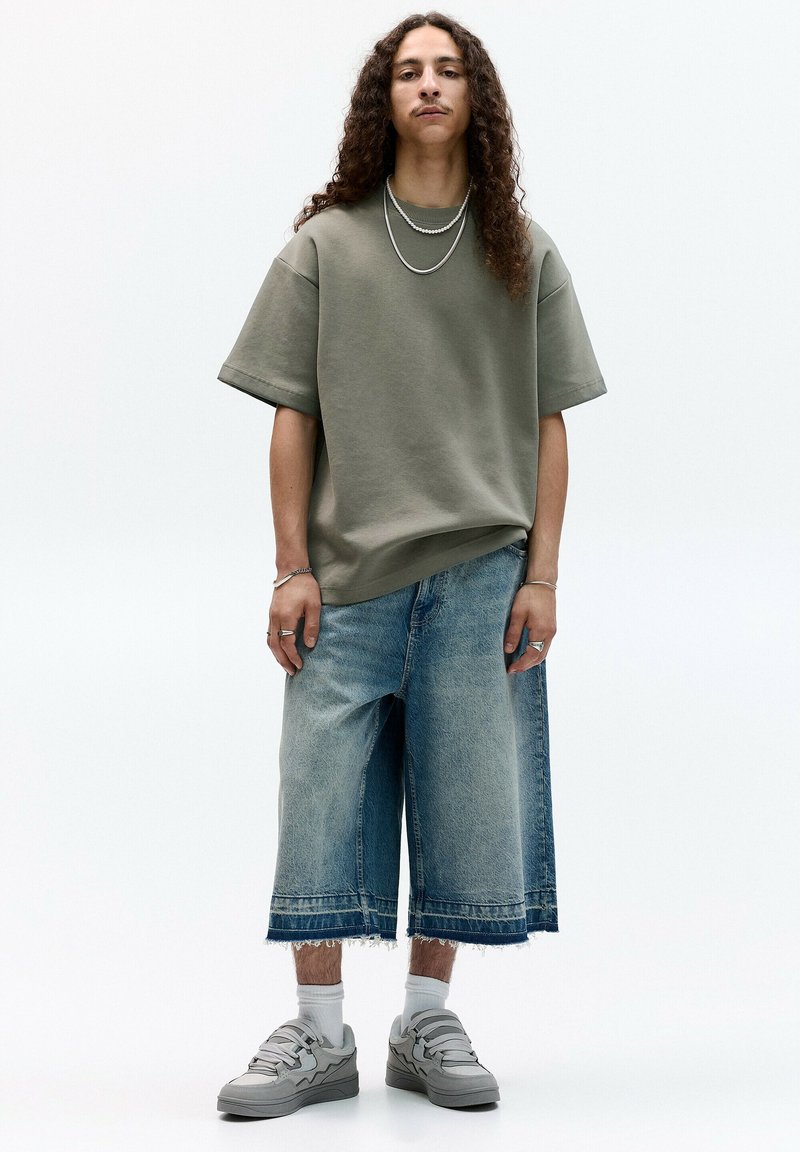 Overdimensioneret olivengrøn t-shirt, lyseblå denimshorts med frynset kant, grå sneakers med lagdelte stropper, lagdelte halskæder.