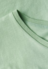 Jasnozielony materiał koszulki z gładką fakturą, z okrągłym dekoltem w prążki i subtelnymi detalami szewnymi w dopasowanym kolorze.