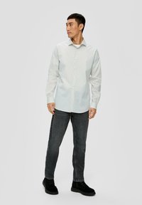 Weißes Hemd mit kleinen grünen Punkten, langen Ärmeln und klassischem Kragen. Kombiniert mit dunkelgrauen Jeans und schwarzen Schuhen.