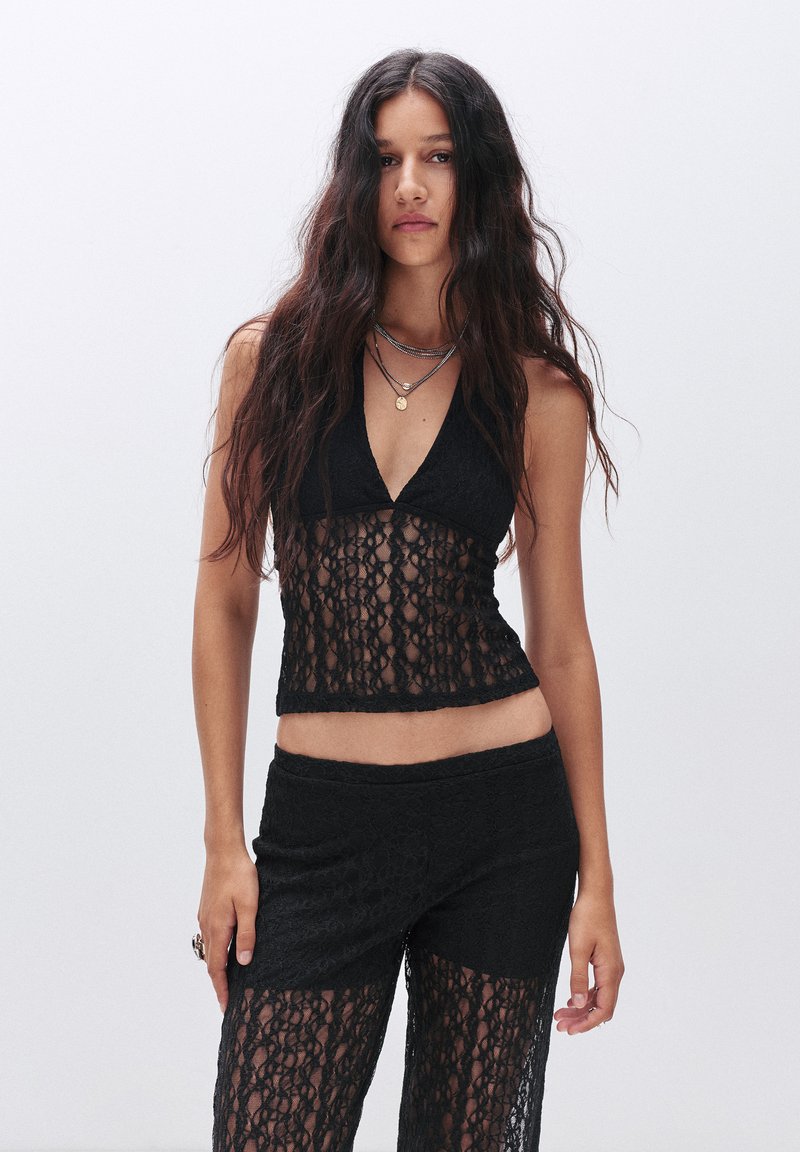 Top halter in pizzo nero con scollatura a V, caratterizzato da intricati motivi floreali e una texture trasparente, abbinato a pantaloni in pizzo coordinati.