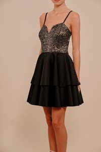 Žena u bezručnom crnom haljini s uskim bodicem ukrašenim šljokicama i slojevitom raširenom suknjom, stoji pred jednostavnom bež pozadinom.