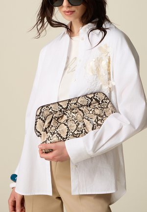 Femme portant une chemise blanche et un pantalon beige, tenant une grande pochette motif peau de serpent sur un fond neutre.
