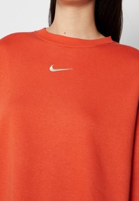 Pomarańczowa bluza z okrągłym dekoltem, z małym złotym logo Nike na piersi. Wykonana z miękkiego, teksturowanego materiału.