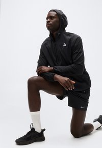 Άνδρας που φοράει μαύρο μπουφάν με κουκούλα και σορτς γονατίζει στο ένα γόνατο, κοιτάζοντας μπροστά μπροστά σε απλό φωτεινό φόντο.