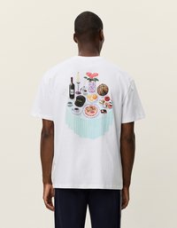 Vit bomulls t-shirt med en färgglad grafisk design av ett bord dukat med matartiklar, blommor och drycker på baksidan.