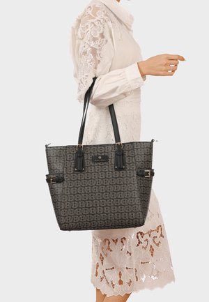 Donna che indossa un vestito bianco in pizzo e porta una borsa tote nera con motivo, dotata di due spalline e dettagli a fibbia.