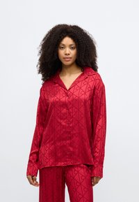 Set de pijamale roșii din satin cu un model geometric. Cămașa cu mâneci lungi are guler și închidere cu nasturi. Pantalonii au același design.