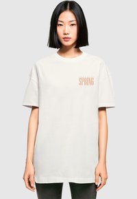 Hvid bomulds T-shirt med korte ærmer, forsynet med et "Tropical Spring" print i lys orange på venstre brystområde.