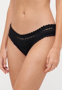 Melna tamborēta bikini apakša ar viļņveida malām, teksturētu rakstu un zemu jostasvietu, kas parāda sarežģītu meistarību un dizaina detaļas.
