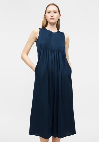 Γυναίκα που φοράει midi φόρεμα χωρίς μανίκια σε σκούρο μπλε χρώμα με πλισέ μπούστο και τσέπες, στέκεται μπροστά σε ένα απλό φωτεινό φόντο.
