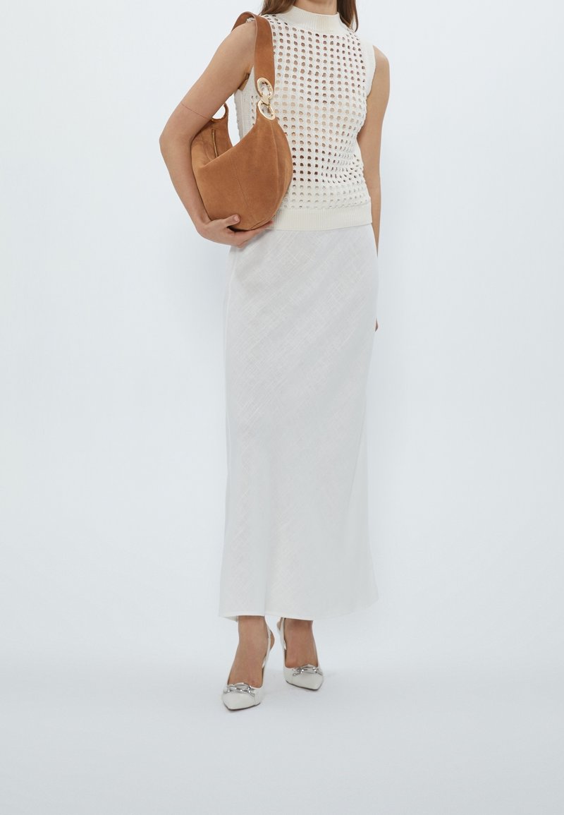 Femme portant un haut blanc en crochet sans manches, une longue jupe blanche, des chaussures blanches pointues avec des nœuds, tenant un sac à bandoulière en daim marron.