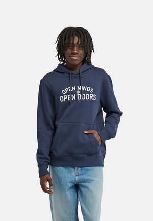 Personne debout, souriante, portant un hoodie bleu marine avec le texte "Open Minds Open Doors" et un jean bleu clair sur un fond clair uni.