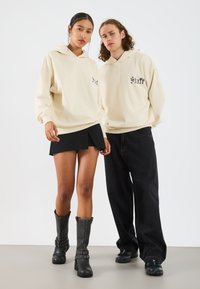 Felpe beige con testo "Stuff" nero, indossate con una gonna nera a pieghe e jeans neri larghi. Stivali neri fin sotto il ginocchio con dettagli borchiati.