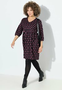 Musta mekko, jossa on vaaleanpunainen geometrinen kuvio, V-pääntie, kolme neljäsosaa hihat, rypytetty vyötärö, käytetään mustien sukkahousujen ja nilkkasaapikkaiden kanssa.