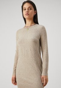 Bežs adīts svārku kleita ar garām piedurknēm, ribotām aprocēm un apakšmalu, ar smalku faktūru un apaļu kakla izgriezumu. Akcentēta ar zaļu kulonu.