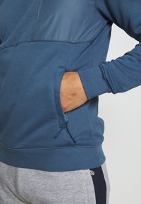 Felpa sportiva blu con un pannello superiore liscio e polsini a coste. Presenta una tasca laterale con zip e una texture morbida.