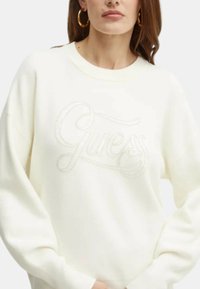 Felpa bianca con un evidente logo ricamato "Guess" sul davanti. Il design include maniche lunghe e colletto rotondo.