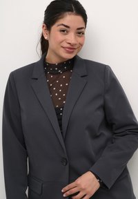 Šedý blazer s jedním knoflíkem, ostrými klopami a dvěma předními kapsami, vrstvený přes černý top s puntíky z jemné síťoviny. Hladká texturka látky.