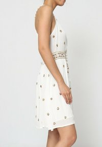 Robe blanche avec une texture plissée, des accents brodés en or et une taille cintrée. Présente des bretelles fines et une silhouette évasée.