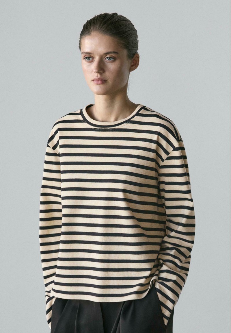 Junge Frau mit zurückgekämmtem Haar trägt ein beige-schwarz gestreiftes Langarmshirt und steht mit den Händen in den Taschen vor einem schlichten Hintergrund.