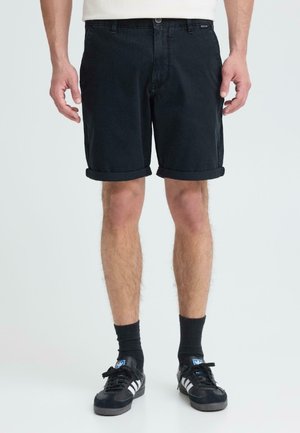 Homme portant un short noir retroussé, des chaussettes noires et des baskets Adidas noires, debout devant un fond clair uni.