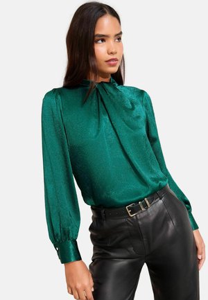 Smaragdgrüne Bluse mit hohem Kragen, langen Puffärmel und einem glänzenden, strukturierten Stoff; kombiniert mit hoch taillierten schwarzen Lederhosen.
