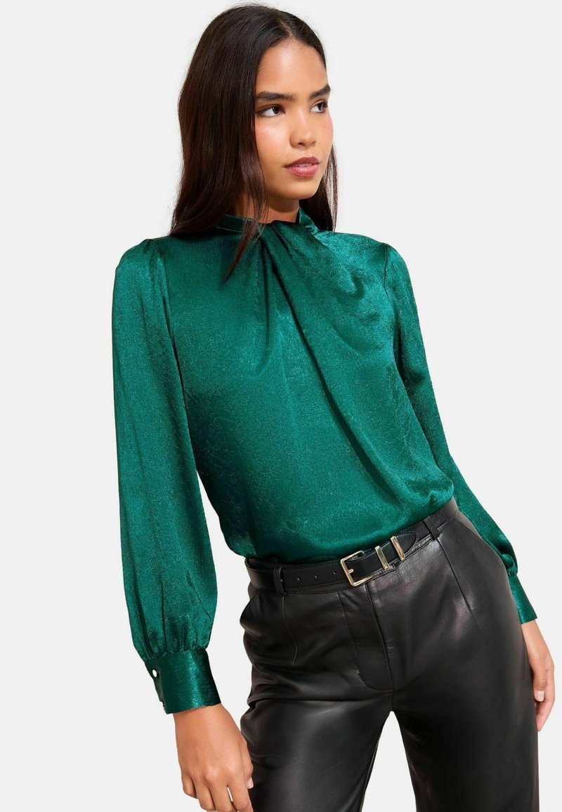 Smaragdrohe bluus kõrge kaelusega, pikkade puhvärvidega ja läikiva, tekstureeritud kangaga; koos kõrgelt taljest mustade nahkpükstega.