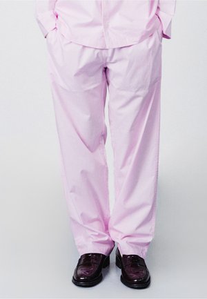 Henkilö seisoo käsineet taskuissa, pukeutuneena löysiin vaaleanpunaisiin housuihin ja kiiltäviin tummanruskeisiin pukukenkiin yksinkertaisen taustan edessä.